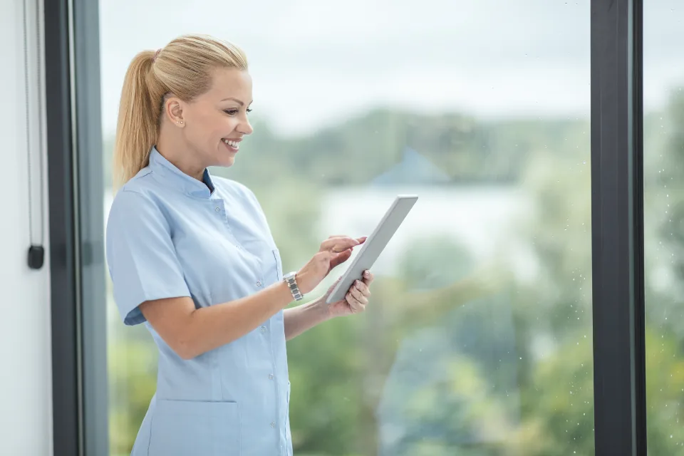 A nurse in scrubs smiling at a tablet beside a large window